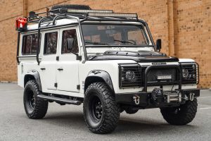 Modified white Land Rover Defender 110 with an external roll cage, roof rack, and front winch