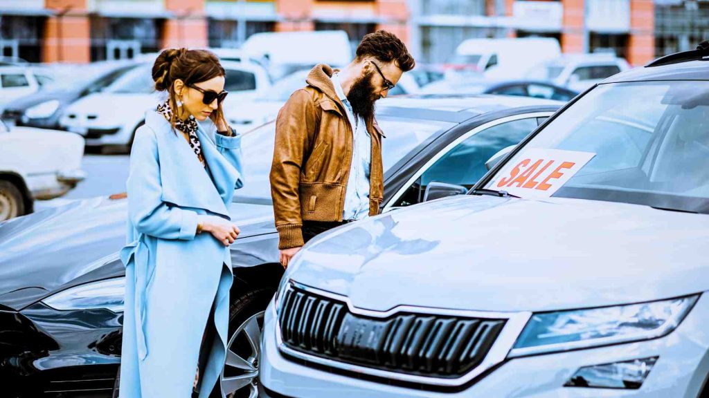 a couple inspecting a used car that has the sign sale on it as if checking a used car at auction