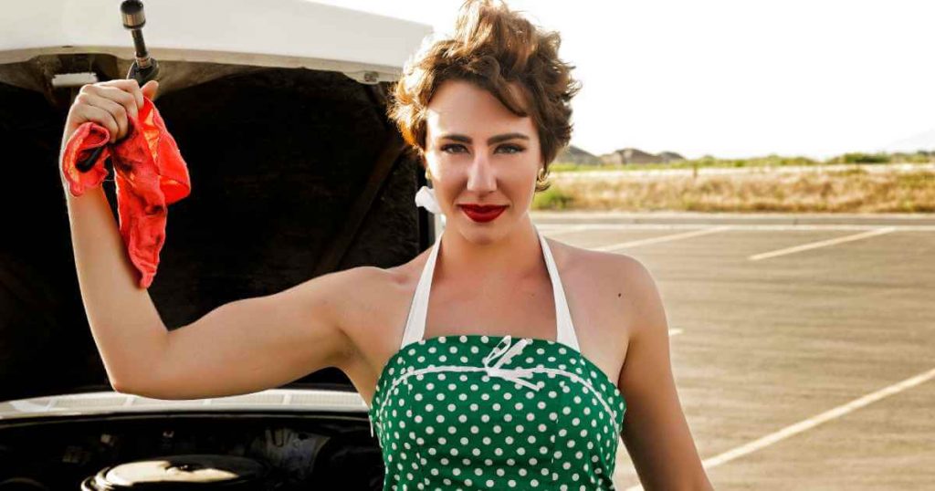 a confident woman in a green dress flexing her arm with an oily rag in her hand and a car with the hood propped up as she performs car repairs herself