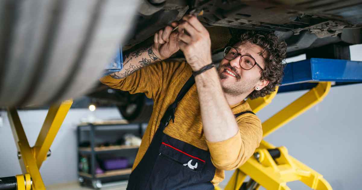 a happy man in a bright yellow shirt working on a car repair with the used car propped in the air