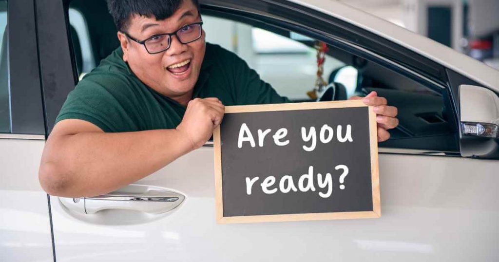 a happy man hanging out of the window of a used car with a board that reads "are you ready" for buy here pay here auto sales