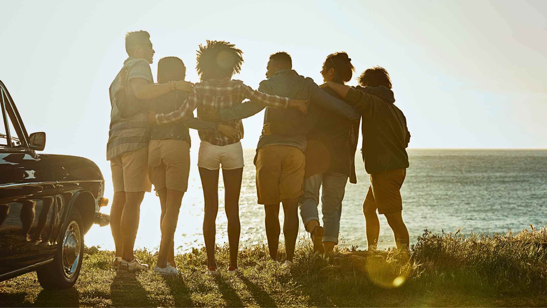 a photo of a group of friends with their arms around each other as they look out over a mountain into the ocean with their car they bought from a buy here pay here near me