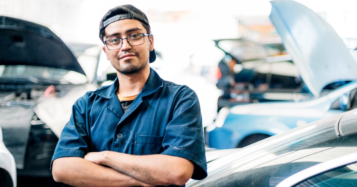 a man in a mechanic's uniform smiling with used cars behind him