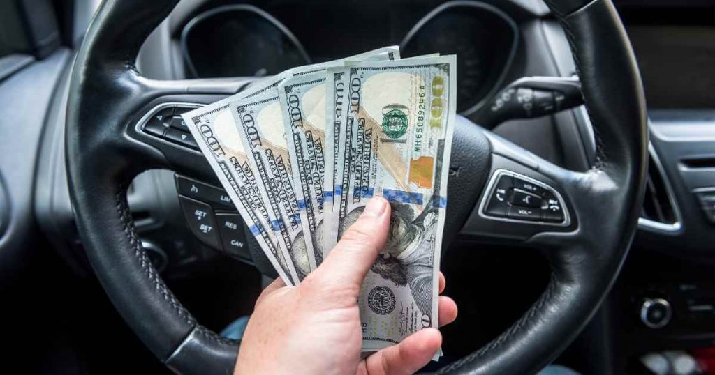 a hand in front of a steering with holding $500 after learning to save money on your car purchase