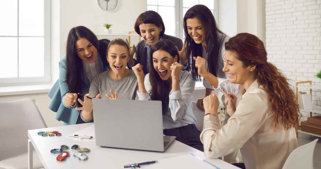 a group of women cheering over something happening on their laptop where toy cars sit next to it showing them getting a real deal on buy here pay here cars